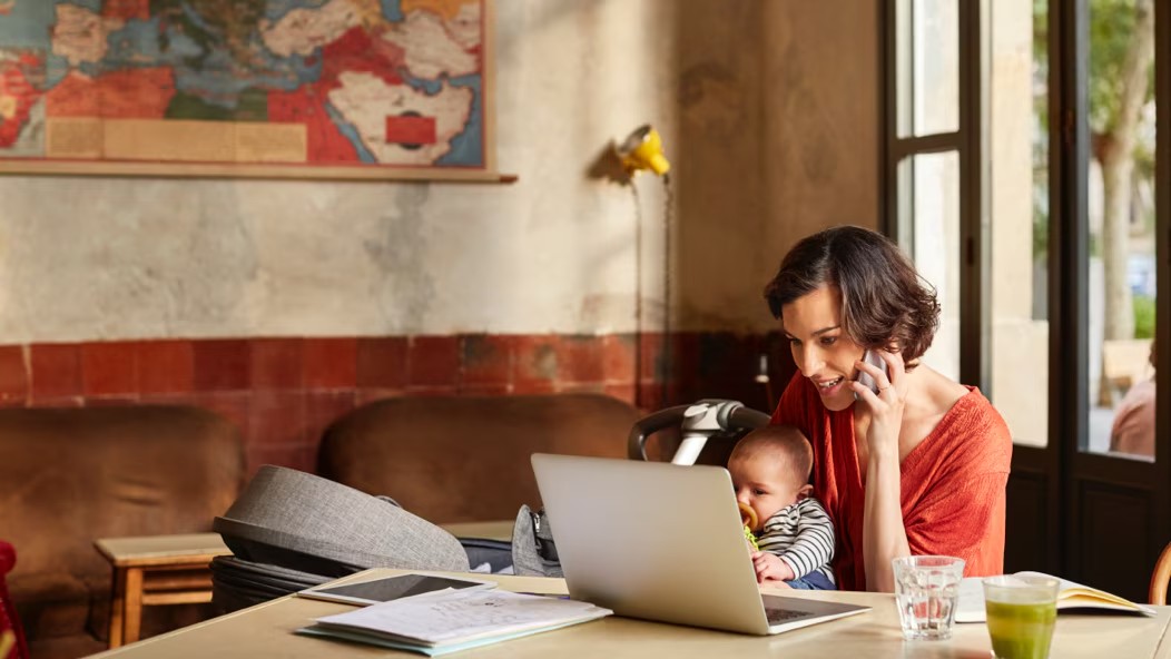 Una persona al teléfono sosteniendo a un niño pequeño y mirando una computadora portátil.